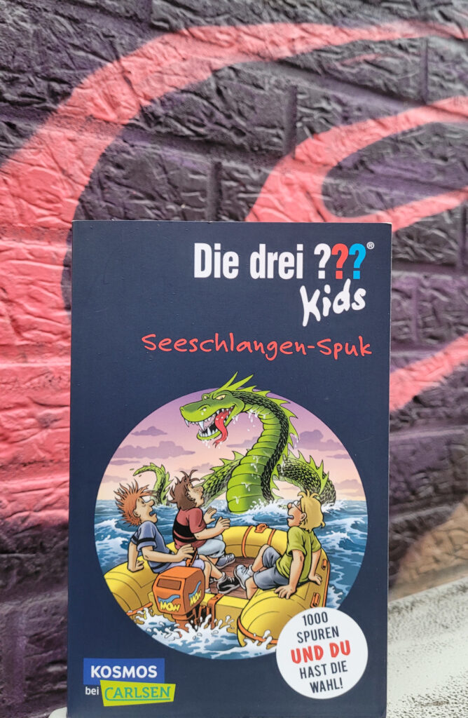Der Kinderkrimi „Die drei ??? Kids und du: Seeschlangen-Spuk“ von Boris Pfeiffer und Harald Juch vor einer Wand mit Grafitti