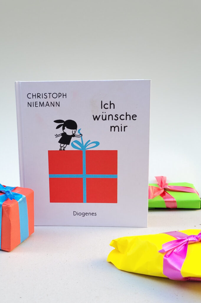 Das Bilderbuch „Ich wünsche mir“ von Christoph Niemann vor weißem Hintergrund umgeben von drei Geschenk-Päckchen