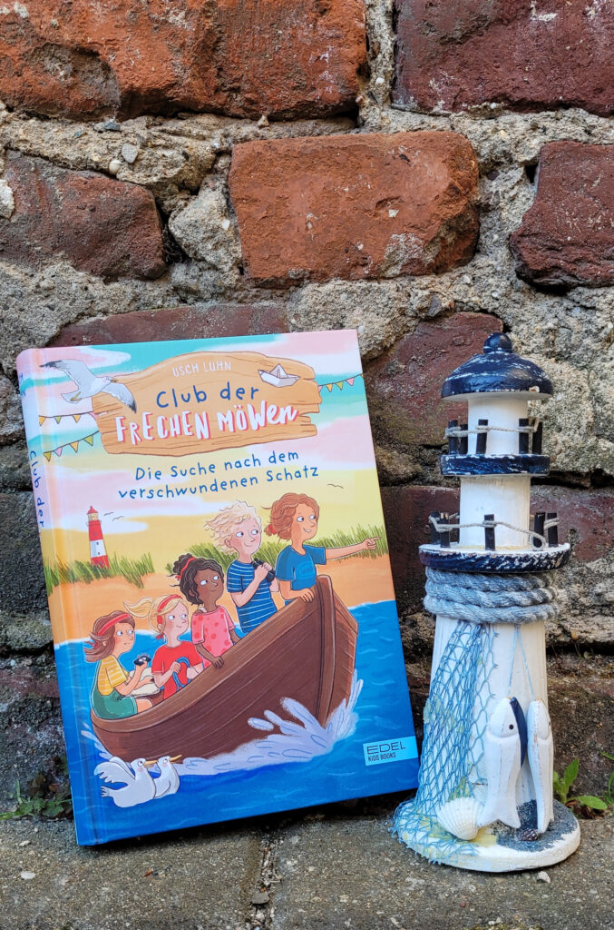 Das Kinderbuch „Club der frechen Möwen: Die Suche nach dem verschwundenen Schatz“ von Usch Luhn vor einer Backsteinmauer neben einem Spielzeug-Leuchtturm