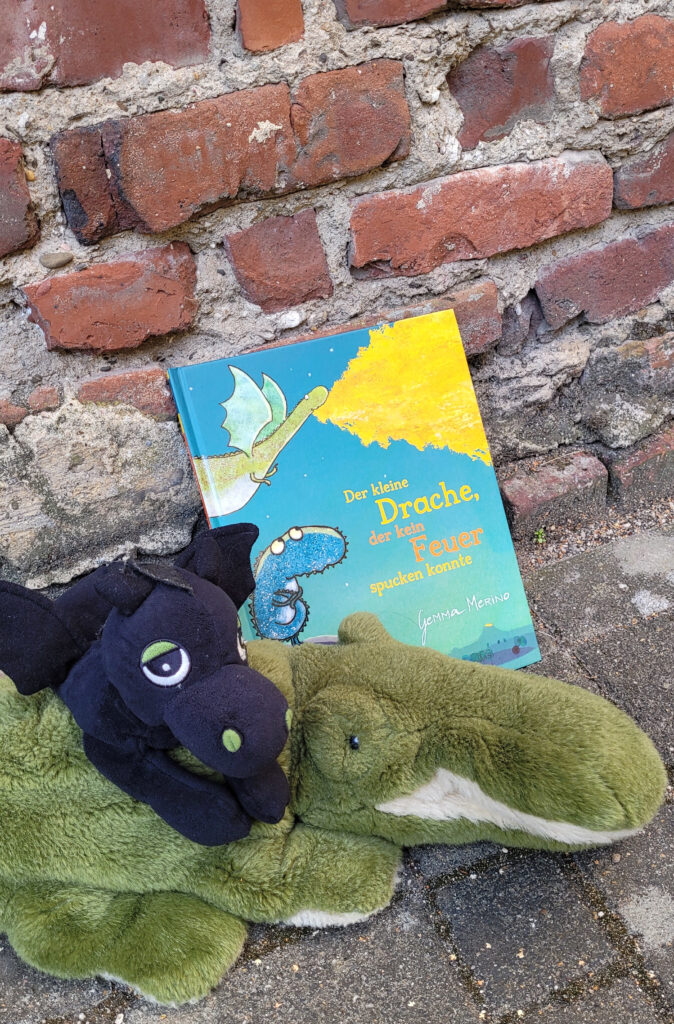 Das Bilderbuch „Der kleine Drache, der kein Feuer spucken konnte“ von Gemma Merino lehnt an einer Backsteinmauer und wird umgeben von einem Drachenkuscheltier und einem Krokodilkuscheltier