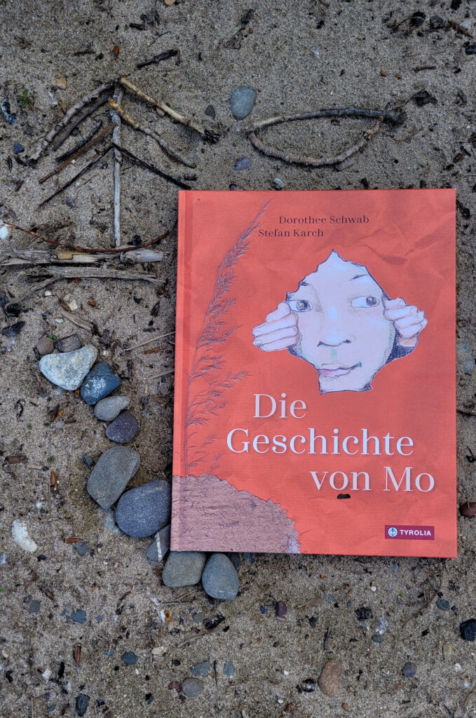 Das Bilderbuch „Die Geschichte von Mo“ von Dorothee Schwab und Stefan Karch auf Sand liegend, drumherum Steine und kleine Äste als Bild angeordnet