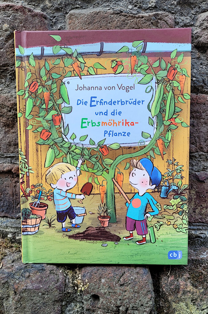 Das Kinderbuch „Die Erfinderbrüder und die Erbsmöhrika-Pflanze“ von Johanna von Vogel vor einer alten Backsteinmauer