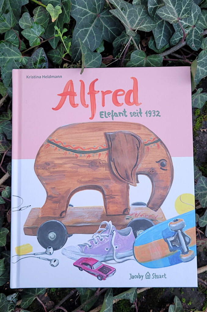 Das Kinderbuch „Alfred - Elefant seit 1932“ von Kristina Heldmann liegt auf einer Backsteinmauer von Efeu umgeben