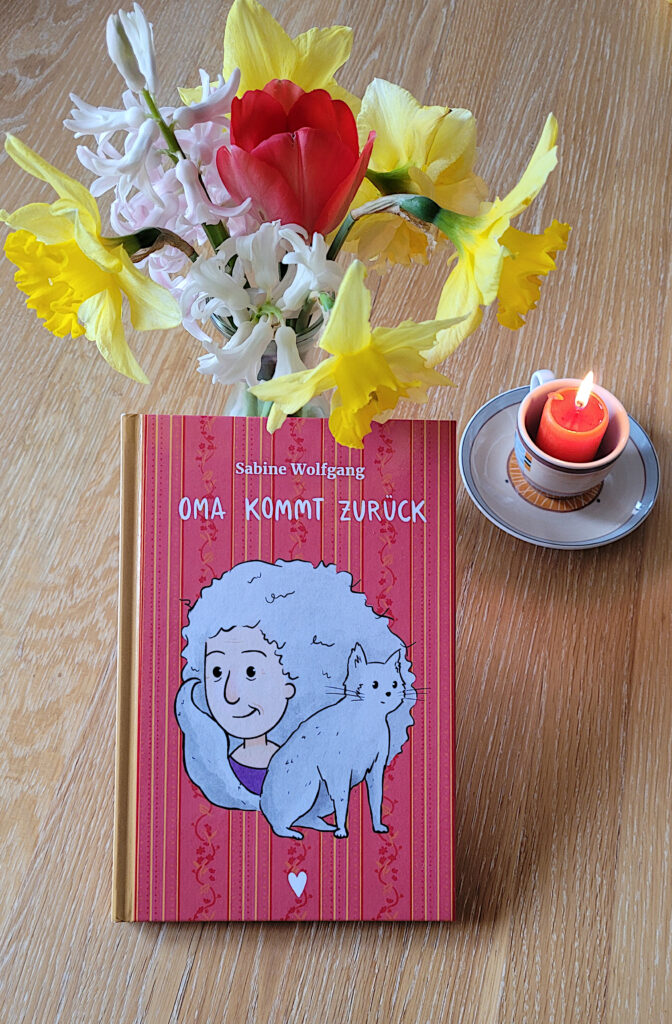 Das Kinderbuch „Oma kommt zurück“ von Sabine Wolfgang vor einem Frühlingsblumenstrauß und einer brennenden Kerze in einer kleinen Tasse.