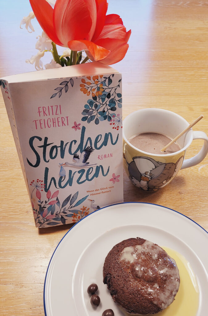 Der Roman „Storchenherzen“ von Friederike Grauf und Mina Teichert auf einem Tisch stehen mit einer roten Tulpe im Hintergrund, einer Tasse heißer Schokolade und einem kleinen Schokokuchen.