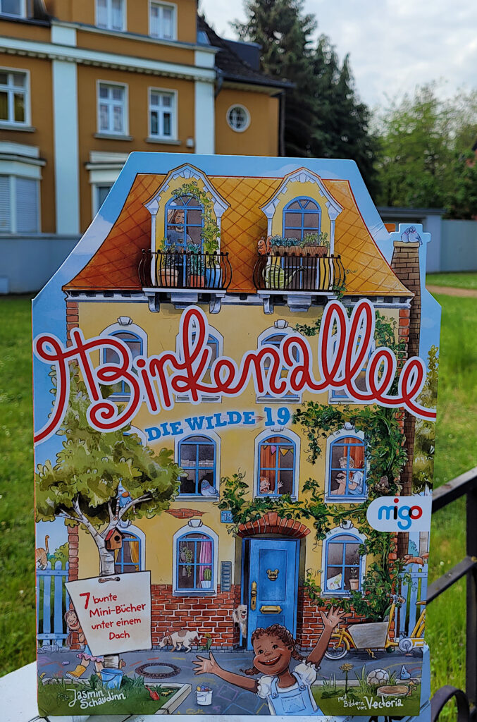 Die Minibuch-Sammlung „Birkenallee. Die wilde 19 - 7 bunte Mini-Bücher unter einem Dach“ von Jasmin Schaudinn, Vectoria und Marc Widmer vor einem orangefarbenen Haus.