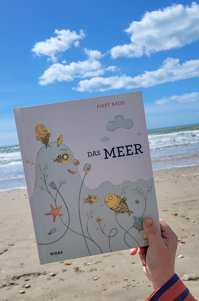 Das Bilderbuch „Das Meer“ von Piret Raud von einer Hand vor Strand und Meer gehalten
