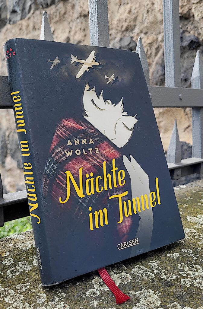 Das Jugendbuch „Nächte im Tunnel“ von Anna Woltz auf einer Mauer vor einem Zaun