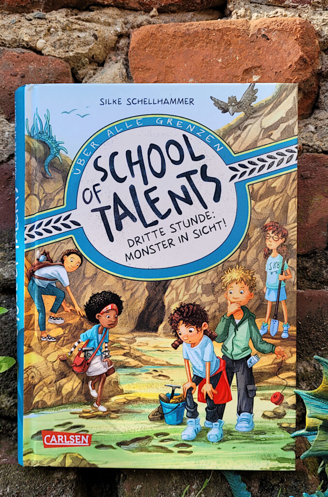 Das Kinderbuch „School of Talents 3: Dritte Stunde - Monster in Sicht!“ von Silke Schellhammer vor einer roten Backsteinmauer