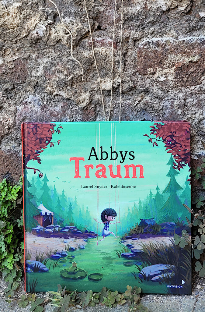 Das Bilderbuch „Abbys Traum“ von Laurel Snyder und Kaleidoscube vor einer Backsteinmauer stehend, von Schnüren gehalten.