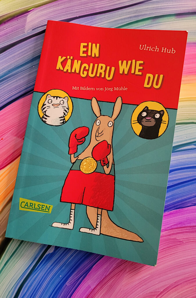 Das Kinderbuch „Ein Känguru wie du“ von Ulrich Hub auf einem Regenbogen-Bild liegend
