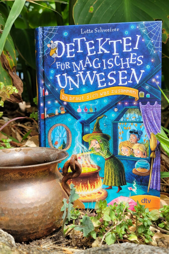 Das Kinderbuch „Detektei für magisches Unwesen: Da braut sich was zusammen“ von Lotte Schweizer vor Pflanzen stehend. Davor steht ein kupferner Hexenkessel.