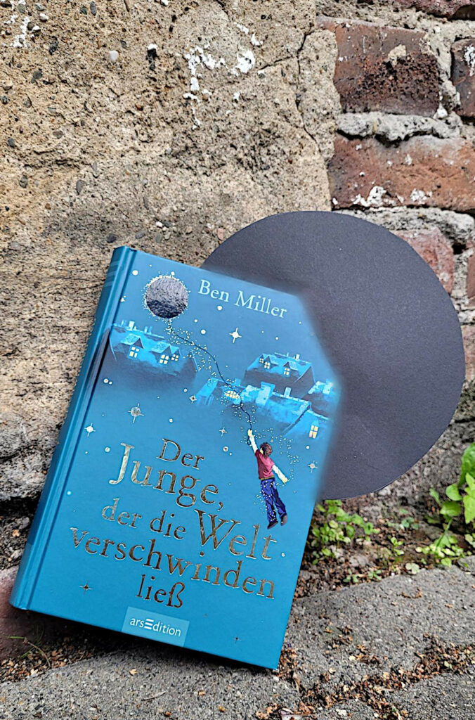 Das Kinderbuch „Der Junge, der die Welt verschwinden ließ“ von Ben Miller lehnt an einer Backsteinmauer und wird von einem Schwarzen Loch eingesaugt
