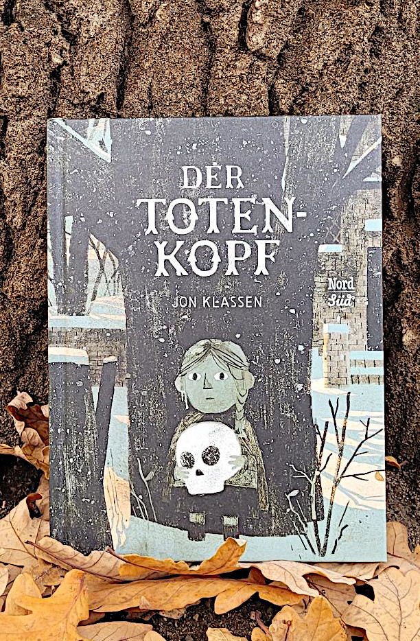 Das Bilderbuch „Der Totenkopf“ von Jon Klassen in Herbstlaub vor einem Baum stehend