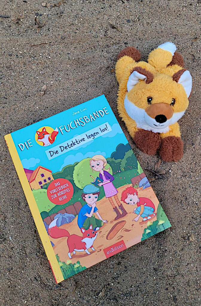 Das Kinderbuch „Die Fuchsbande: Die Detektive legen los!“ von Jana Lini und Tessa Rath liegt in Sandkastensand. Daneben liegt ein Kuscheltierfuchs.