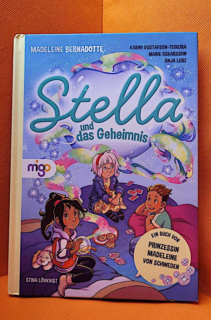 Das Kinderbuch „Stella und das Geheimnis“ von Madeleine Bernadotte, Karini Gustafson-Teixeira, Marie Oskarsson und Stina Lövkvist vor einem orangefarbenen Hintergrund