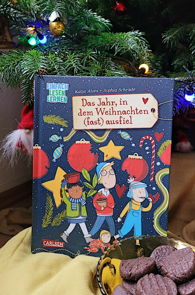 Das Erstlesebuch „Das Jahr, in dem Weihnachten (fast) ausfiel“ von Katja Alves und Sophia Schrade vor einem Weihnachsbaum mit einer Schlae Schokoladenplätzchen