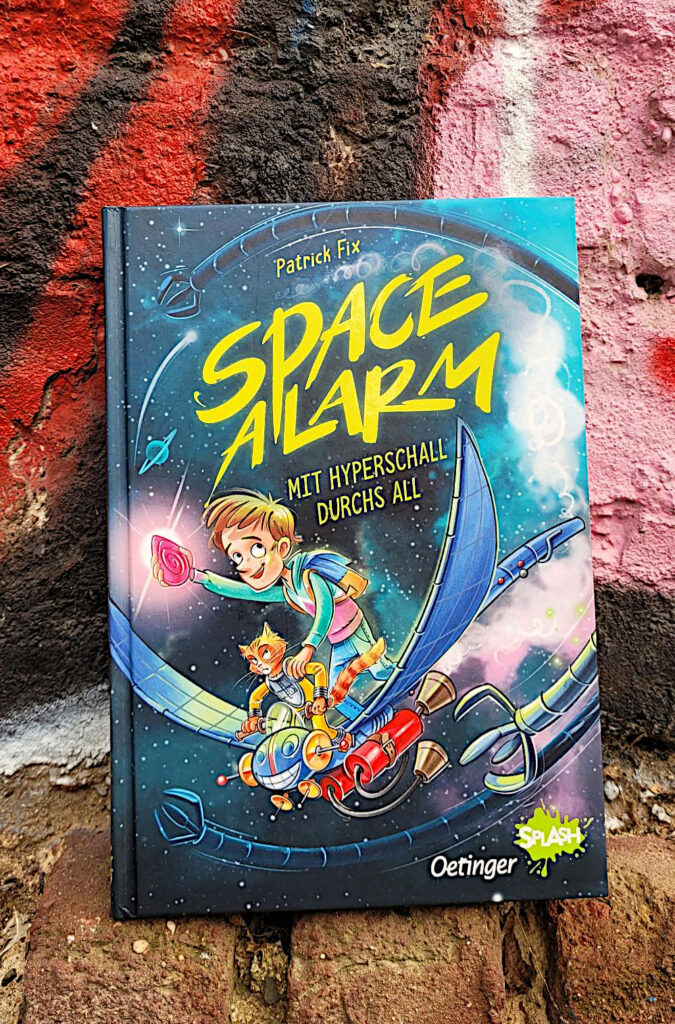 Das Kinderbuch „Space Alarm: Mit Hyperschall durchs All“ von Patrick Fix vor einer mit Graffiti besprühten Backsteinmauer