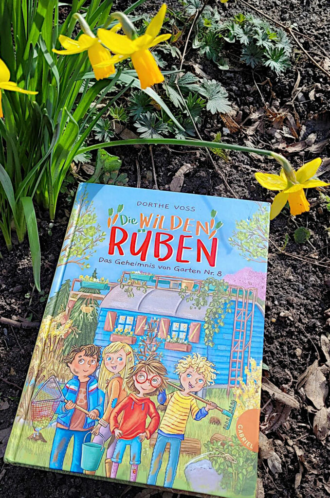 Das Kinderbuch „Die Wilden Rüben: Das Geheimnis von Garten Nr. 8“ von Dorthe Voss auf Erde liegend von Osterglöckchen umrahmt