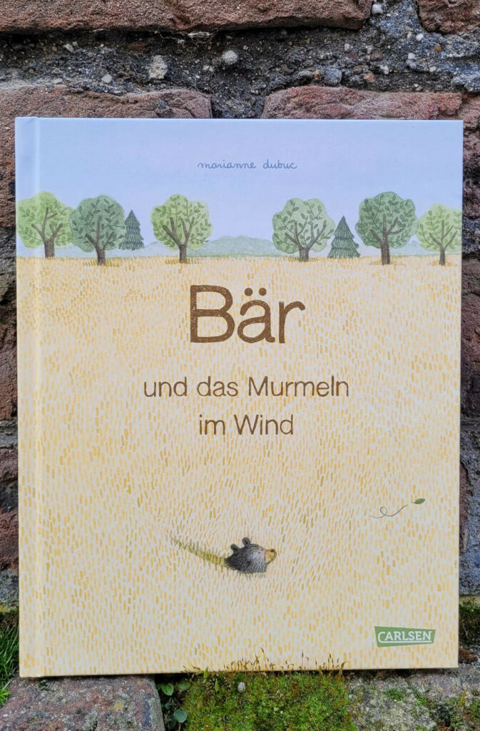 Das Bilderbuch „Bär und das Murmeln im Wind“ von Marianne Dubuc vor einer Roten Backsteinmauer