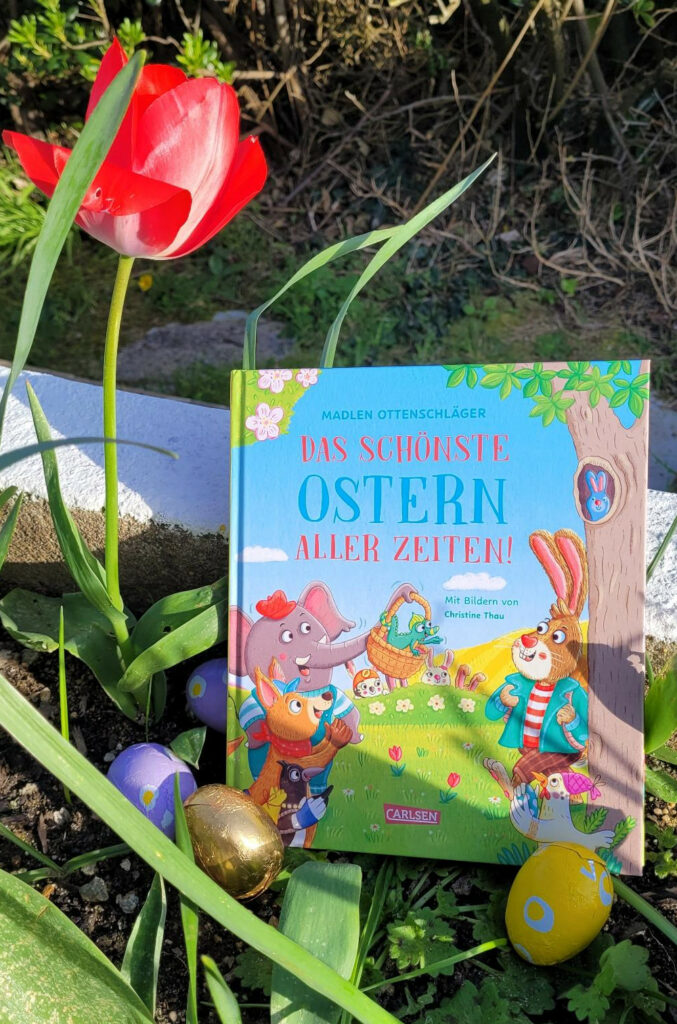 Das Kinderbuch „Das schönste Ostern aller Zeiten!“ von Madlen Ottenschläger und Christine Thau in einbem Blumenbeet neben einer blühenden roten Tulpe und drei verpackte Schoko-Ostereier
