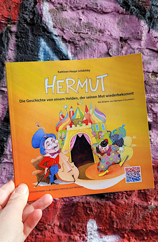 Das Kinderbuch „Hermut: Die Geschichte von einem Helden, der seinen Mut wiederbekommt“ von Kathleen Haupt-Schibilsky und Barbara Schumann vor einer bunt besprühten Backsteinmauer