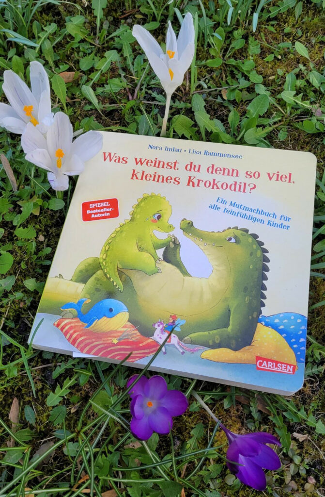 Das Pappbilderbuch „Was weinst du denn so viel, kleines Krokodil?“ von Nora Imlau und Lisa Rammensee auf einer blühenden Frühlingswiese