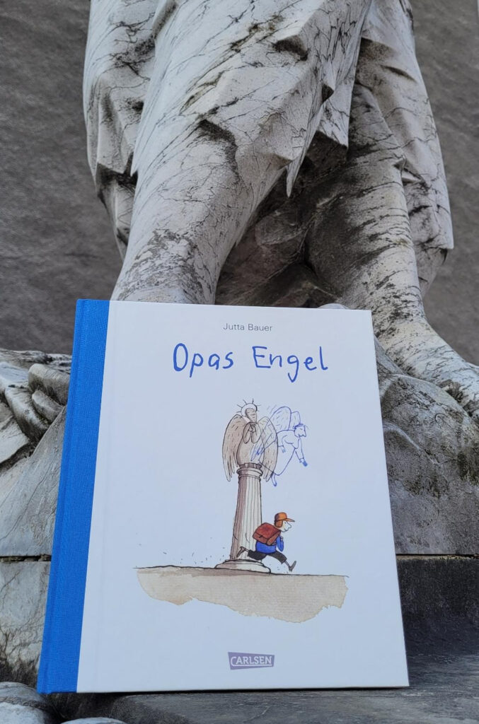 Die Jubiläumsausgabe des Bilderbuches „Opas Engel“ von Jutta Bauer vor den Beinen einer Marmorstatue