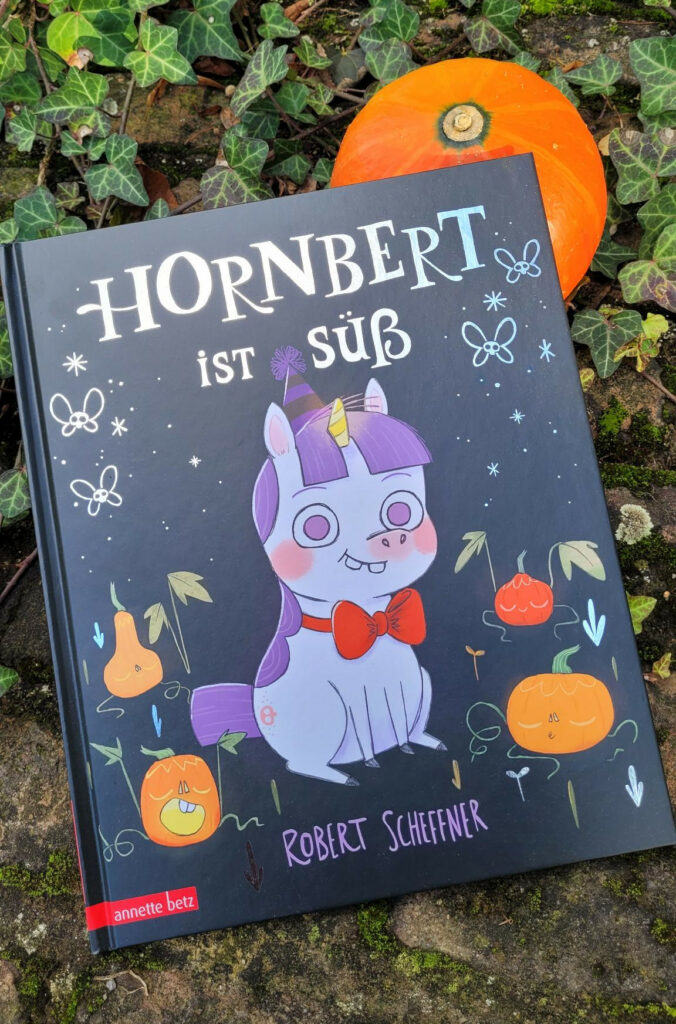 Das Bilderbuch „Hornbert ist süß“ von Robert Scheffner halb auf einem kleinen Kübis liegend, umgeben von Efeu