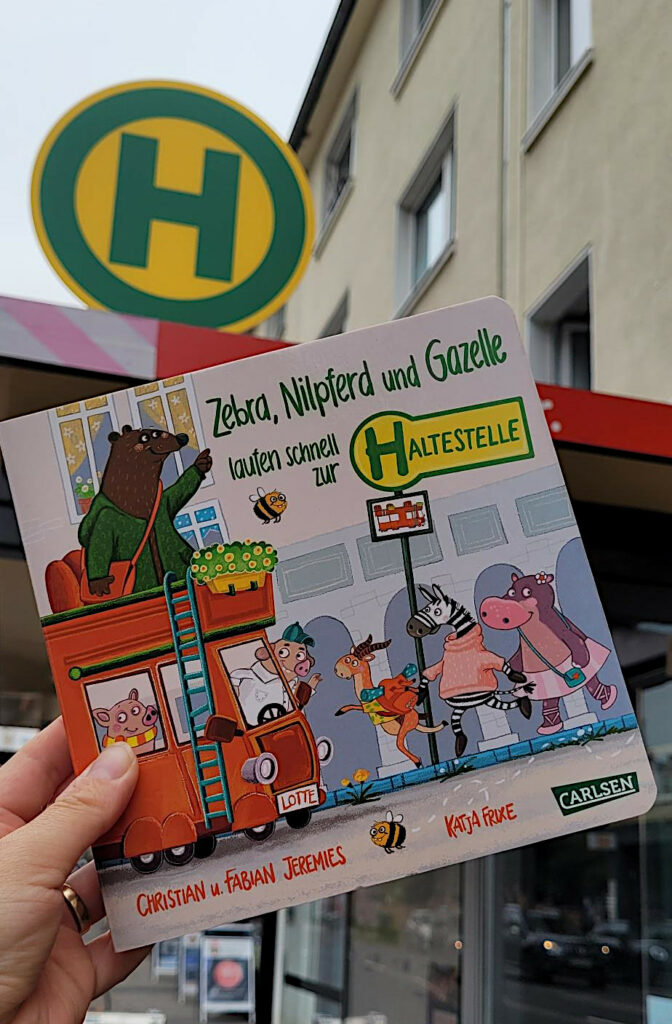 Das Pappbilderbuch „Zebra, Nilpferd und Gazelle laufen schnell zur ... Haltestelle“ von Katja Frixe, Christian Jeremies und Fabian Jeremies vor einer Kölner Bushaltestelle