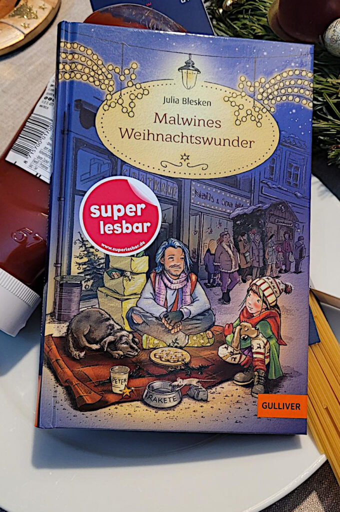 Das Kinderbuch „Malwines Weihnachtswunder“ von Julia Blesken auf einer Flasche Ketchup und Nudeln liegend. Im Hintergrund sieht man Tannengrün und Weihnachtsschmuck