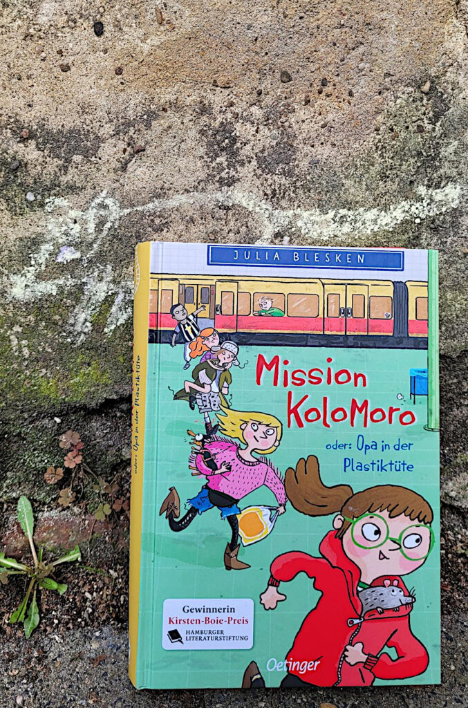 Das Kinderbuch „Mission Kolomoro oder: Opa in der Plastiktüte“ von Julia Blesken vor einer alten Mauer auf die eine Kreidemaus gekritzelt wurde.