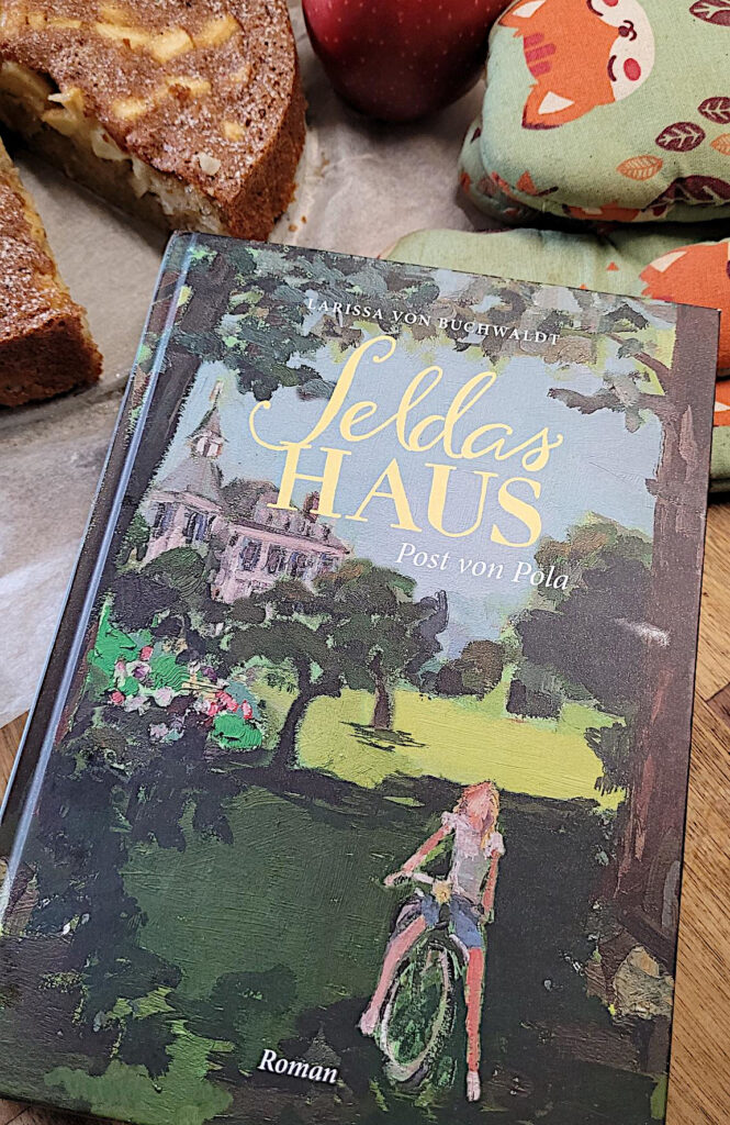 Der Roman „Seldas Haus: Post von Pola“ von Larissa von Buchwaldt auf einerm Tisch liegend. Im Hintergrund sieht man Ofenhandschuhe und einen angeschnittenen Apfelkuchen.