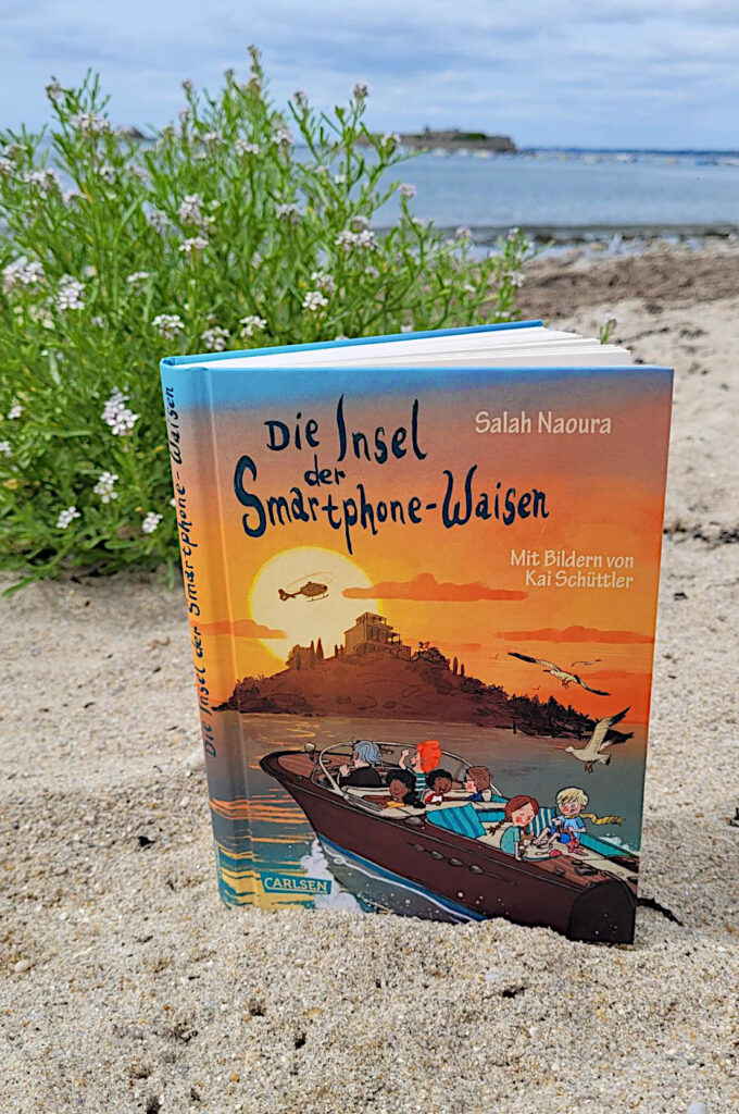 Das Kinderbuch „Die Insel der Smartphone-Waisen“ steht auf einem Sandstrand, im Hintergrund eine Strandpflanze und eine Festung im Meer.