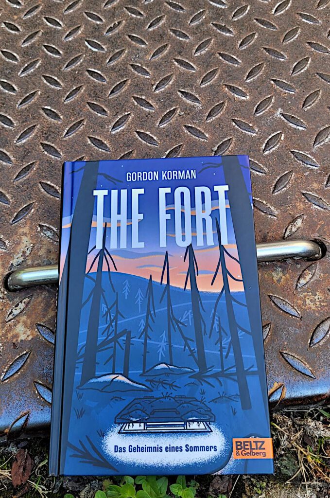 Der Jugendroman „The Fort: Das Geheimnis eines Sommers“ von Gordon Korman liegt auf einer metallenen Bodenklappe.
